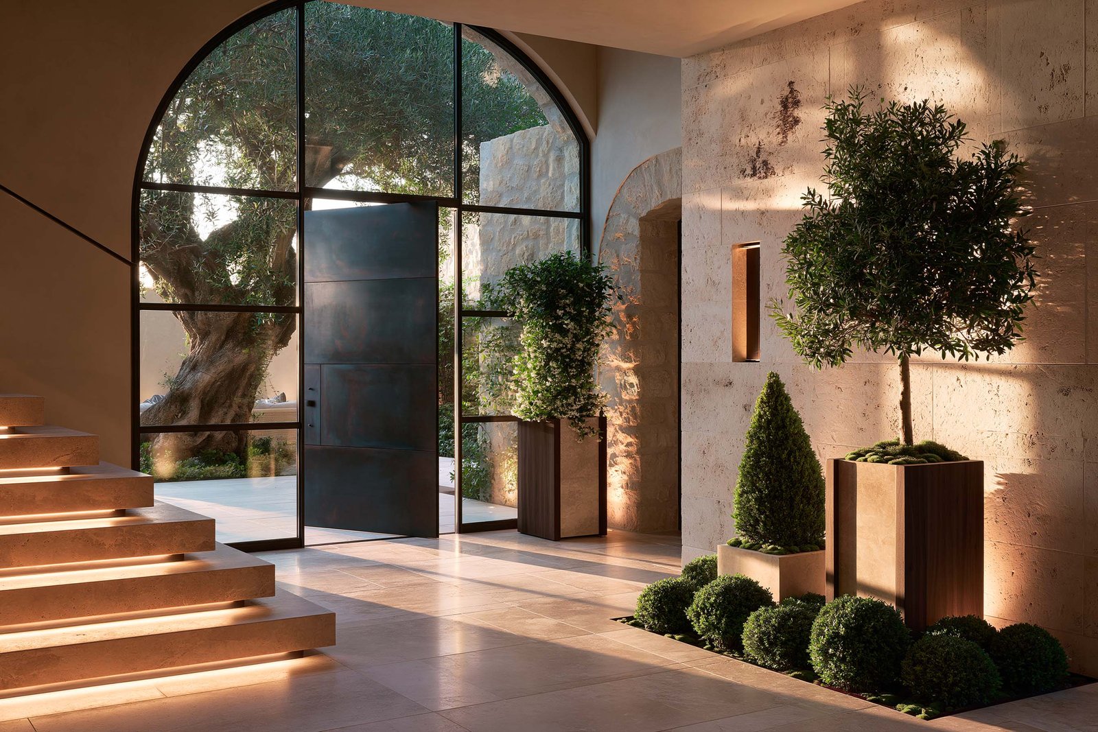 Seuil — guidance botanique dans l’espace : hall d’une villa contemporaine à Èze au crépuscule, escalier travertin cantilever sous LED 2700K, arche vitrée révélant un Olea europaea centenaire dans la cour intérieure, Olea topiaire en pot travertin, Laurus nobilis en topiaire cone et Buxus sempervirens en topiaire boules en pied de mur, Paracelsus Gardens™