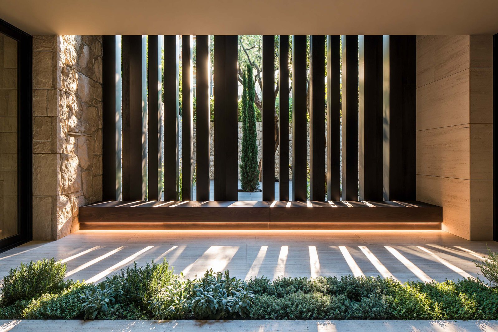 Silhouette structurelle — loggia couverte d’un appartement à Monaco en plein midi, partition SylvaVeil™ en lames verticales d’Acciaio Brunito filtrant la lumière en contre-jour, banc en noyer fumé et petit jardin aromatique de Rosmarinus officinalis, Salvia officinalis et Thymus vulgaris, sol travertin clair, Paracelsus Gardens™