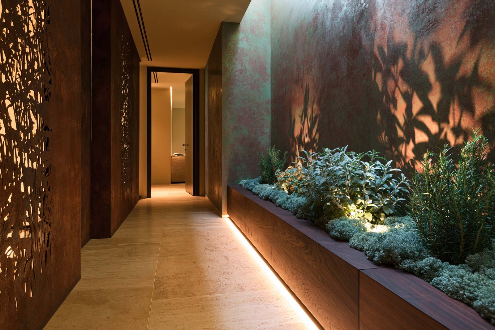 Compression spatiale — couloir intérieur d’une villa à Roquebrune-Cap-Martin en fin de matinée, partition Hortus Velum™ Arcana en Acciaio Brunito à gauche resserrant l’espace, mur en stucco lustro ocre patiné à droite, planter Lignum™ Block en noyer fumé contre la paroi portant Salvia officinalis, Helichrysum italicum et Rosmarinus officinalis, sol travertin clair warm honey, raking light directionnel, Paracelsus Gardens™