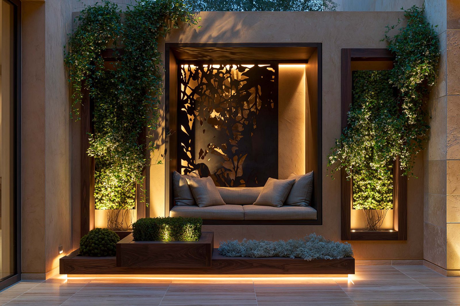 Alcôve intime — alcôve-banc encadrée dans une niche stucco lustro ocre patiné d’une villa à Saint-Paul-de-Vence en fin de journée, banc en noyer fumé sous LED 2700K cachée, partition Hortus Velum™ Arcana en Acciaio Brunito au-dessus projetant ses ombres botaniques sur le mur, plaque verticale de Trachelospermum jasminoides en topiaire encadré flanquant la niche, Buxus sempervirens en topiaire boule compacte au pied, sol travertin clair, Paracelsus Gardens™