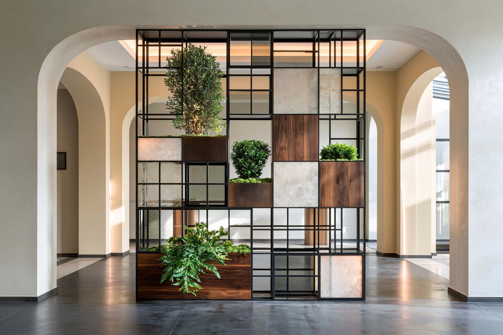 Monaco — atrium d’une résidence contemporaine, Hortus Velum™ Porta freestanding en ossature Acciaio Brunito à profils fins en quinconce non uniforme à densité réduite, panneaux composites en travertin clair, noyer fumé et miroir alternant avec des cellules vides laissant passer la lumière, caissons végétaux intégrés accueillant Olea europaea en topiaire compacte encadrée, Buxus sempervirens en topiaires sculpturales, Asplenium scolopendrium en fougère épée et Dionaea muscipula en accent botanique, arches contemporaines en intonaco crème en arrière-plan, sol micro-béton anthracite poli — Paracelsus Gardens™
