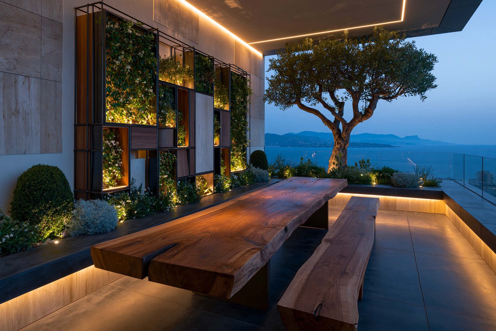 Cap-Ferrat — terrasse penthouse blue hour, Hortus Velum™ Strata mural à composition en quinconce non uniforme dominé par les caissons végétaux, Trachelospermum jasminoides en fleurs blanches étoilées en port encadré et Buxus sempervirens en topiaires, longue table Lignum™ en noyer fumé live-edge taillée dans la masse avec bancs assortis, Olea europaea ancien sculptural en accent extérieur, LED 2700K rasante au plafond et sous les assises, sol en pierre claire ouverte sur la Mer Méditerranée et le relief du Cap d’Antibes — Paracelsus Gardens™