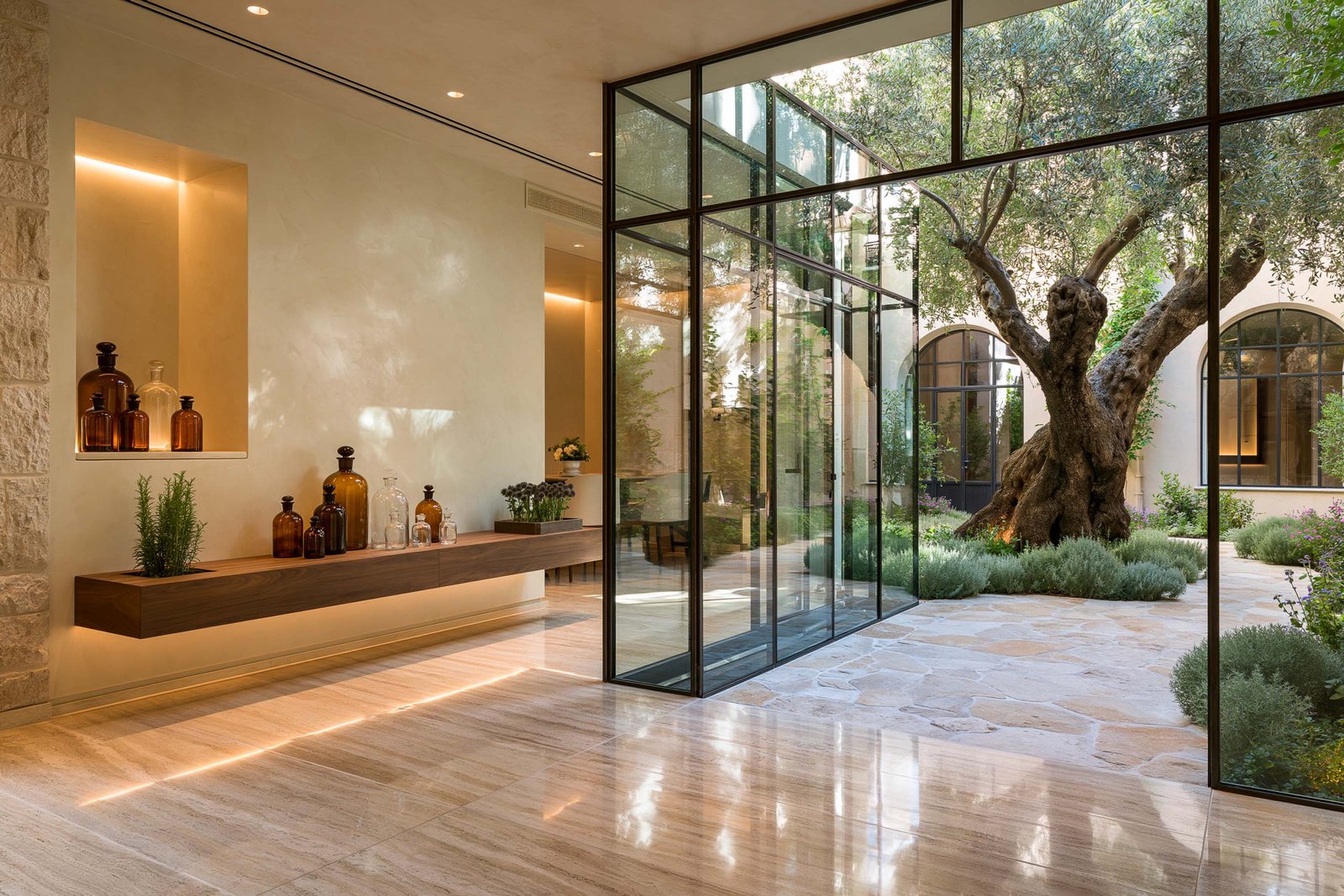 Seuil de transition intérieur-extérieur d’un spa à Beaulieu-sur-Mer, cube vitré en cadre acier noirci, intérieur en travertin clair et console suspendue noyer, cour à olivier mature et masses de lavande visibles.