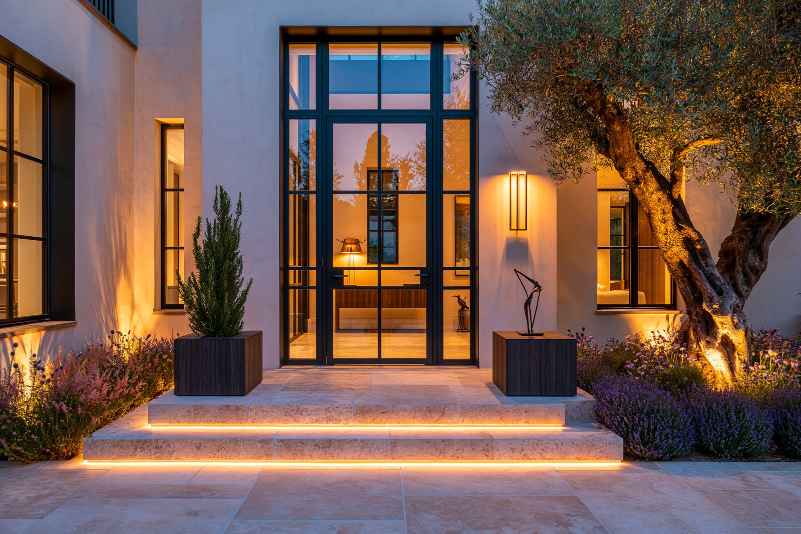 Seuil d’entrée d’une villa à Cap-Ferrat, porte vitrée à cadre rectangulaire acier noirci, jardinières en noyer fumé encadrant le seuil, rétro-éclairage LED 2700K au pied de la plateforme, lumière de fin d’après-midi méditerranéenne.