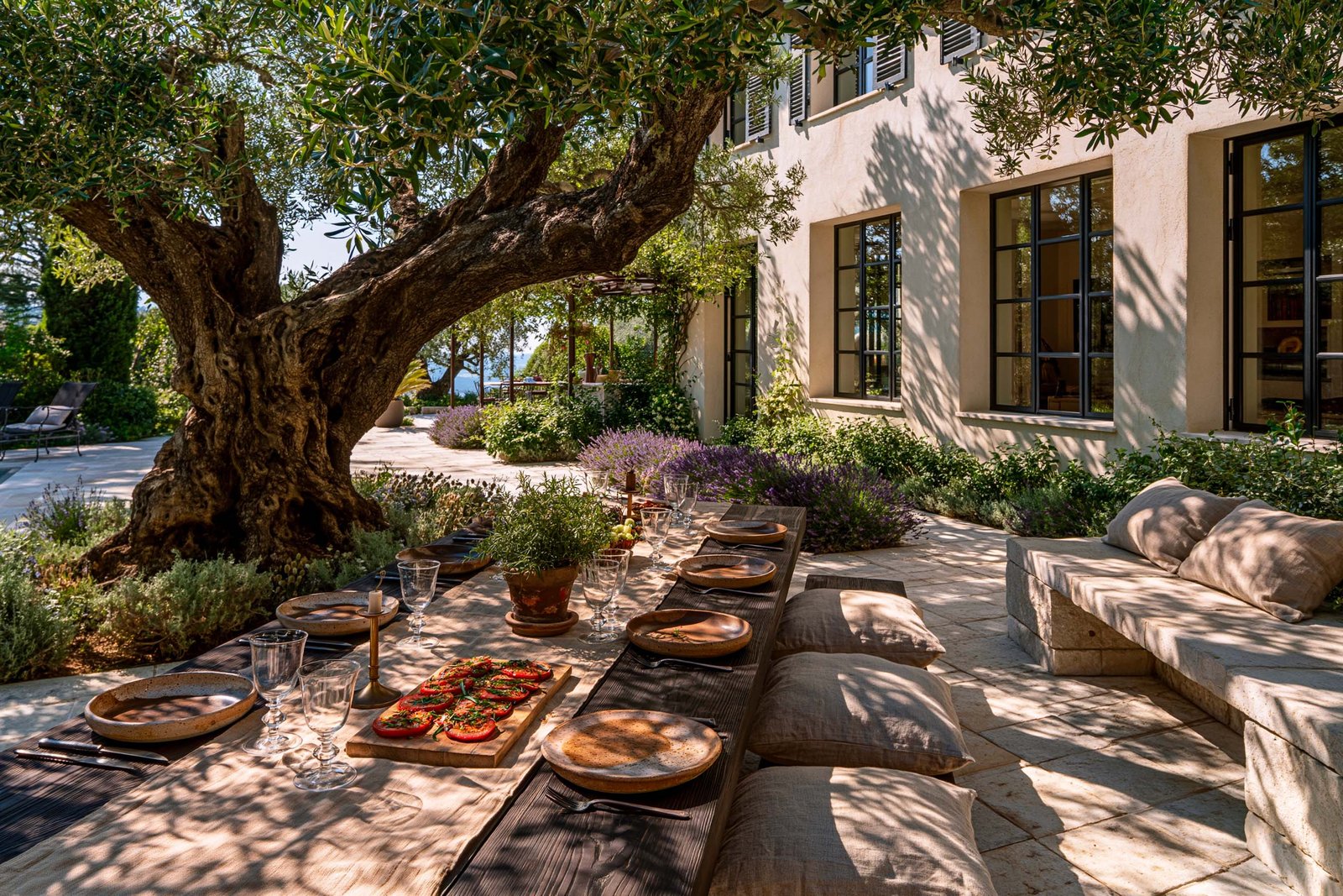 Déjeuner au jardin d’une villa à Saint-Paul-de-Vence, longue table en noyer fumé sous le couvert d’un olivier mature, façade contemporaine en stucco lustro chaud, masses de Lavandula angustifolia et Rosmarinus officinalis en pots terre cuite, dallage pierre claire.