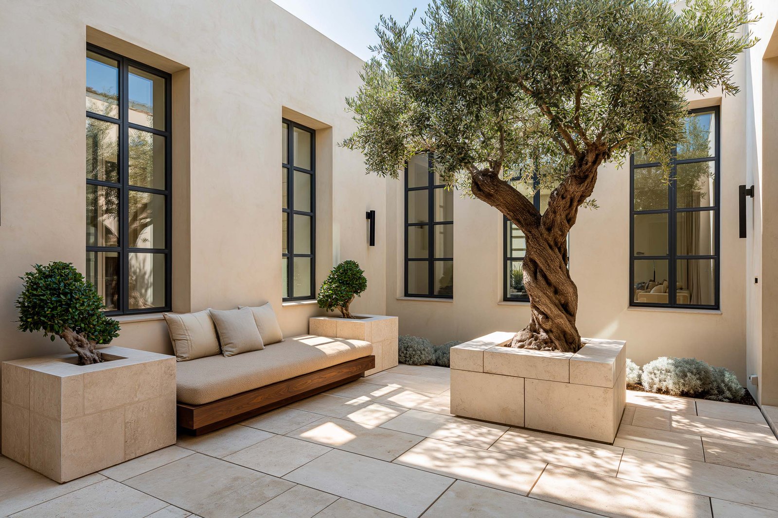 Cour intérieure protégée d’une villa à Saint-Paul-de-Vence, olivier mature dans un bac monolithique en Pietra Serena, banquette flottante en noyer fumé sous une baie vitrée rectangulaire à cadre acier noirci, dallage en travertin clair.