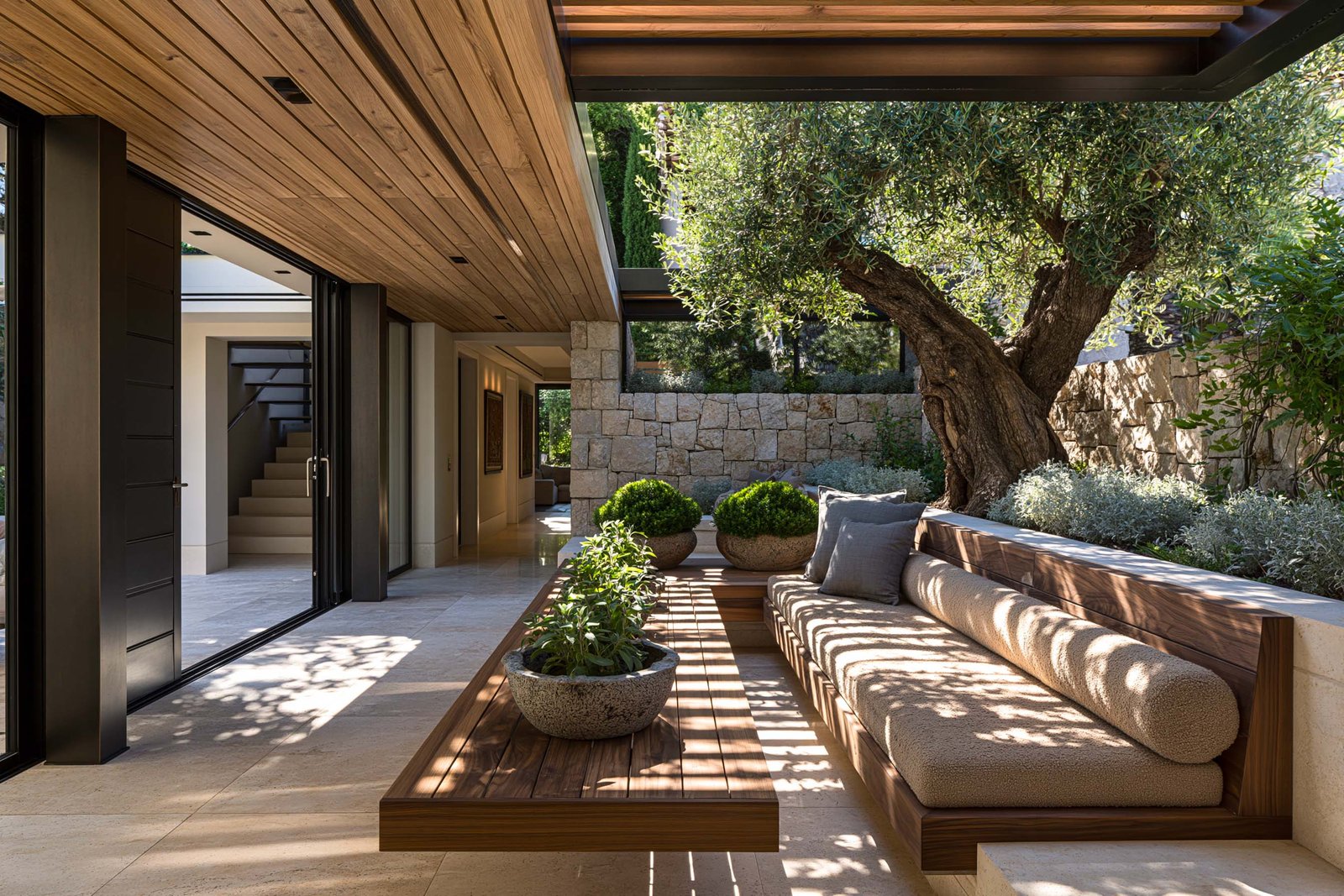 Loggia ombragée d’une villa à Roquebrune-Cap-Martin, plafond en poutres chêne thermotraité propres à linteau droit, banquette intégrée en Pietra Serena avec coussins en lin écru, table en noyer fumé, jardinières d’herbes aromatiques en pots terre cuite.