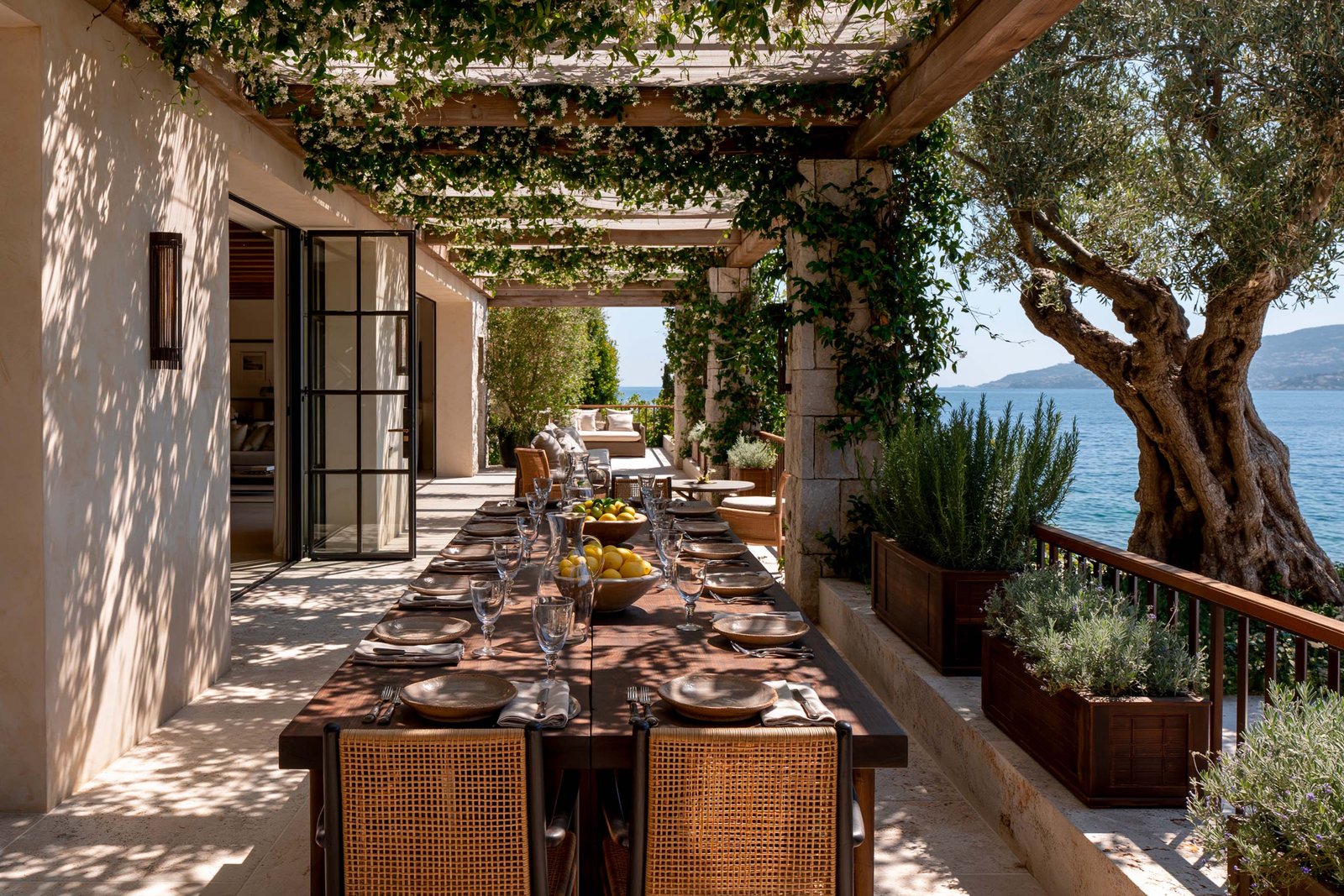 Déjeuner sous pergola d’une villa à Beaulieu-sur-Mer, table en noyer fumé dressée avec citrons et carafe en verre soufflé, pergola en chêne thermotraité avec voile de Trachelospermum jasminoides en port libre, jardinières d’aromatiques, vue sur la mer Méditerranée.