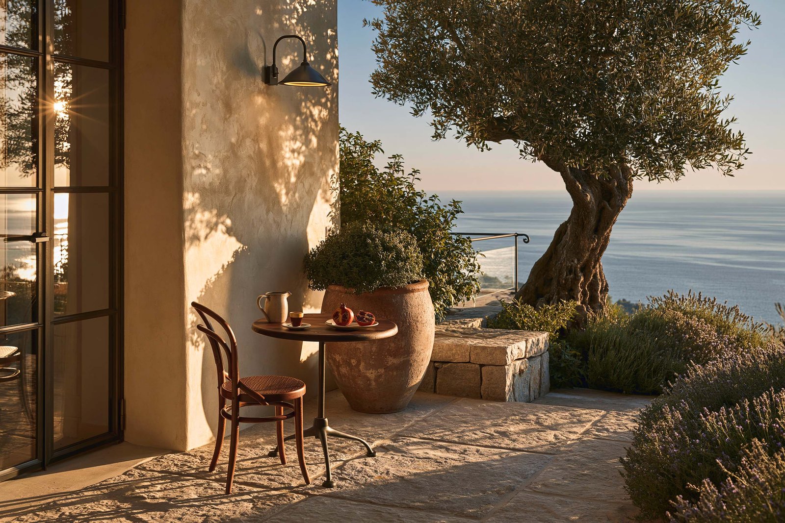 Petit déjeuner du matin sur la terrasse d’une villa à Saint-Paul-de-Vence, petite table ronde en noyer fumé avec tasse en stoneware et corbeille de grenades, façade en stucco lustro chaud, masses de Lavandula angustifolia en pots terre cuite, olivier mature, dallage pierre claire.